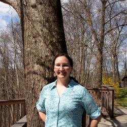 Young woman in a blue button up shirt stands in front of a tree